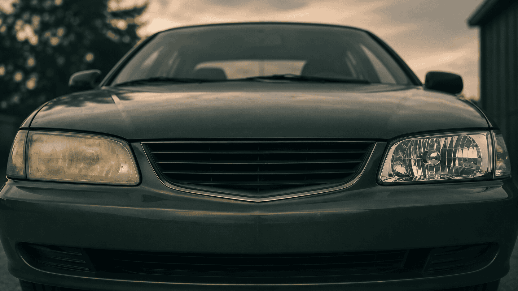 The front of a car at dusk with its two headlights compared, the left one yellowed and cloudy, the right one restored and crystal clear